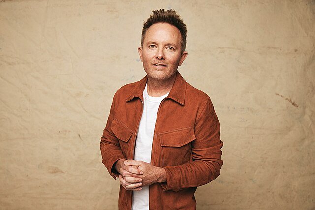 Chris Tomlin in a studio portrait, rust jacket over white tee, hands clasped, neutral beige backdrop.