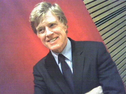 Robert Redford article image: smiling person in a dark suit and tie, three-quarter view against a red background