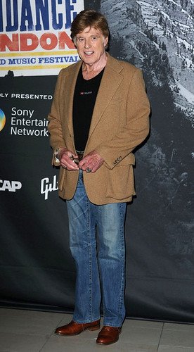 Robert Redford article image: person in a tan blazer, black tee, blue jeans and brown shoes posing at a Sundance London press wall